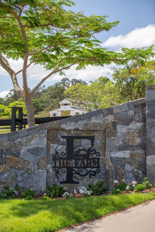 Exterior view of a landscaped property entrance featuring a rough natural-stone boundary wall with a decorative black metal farm sign and gate. Mature leafy trees, lawn, planting, and a partially visible light-coloured house appear behind the wall under a bright sky.