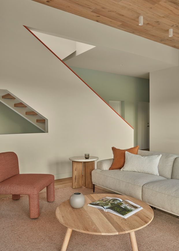 Warm contemporary living room with a sculptural staircase, pale green accent wall, natural timber ceiling and flooring details, neutral upholstered sofa, rust-toned lounge chair, round timber coffee table, and soft textured carpet. The staircase is enclosed by a crisp white balustrade or half-wall with a thin contrasting timber edge.