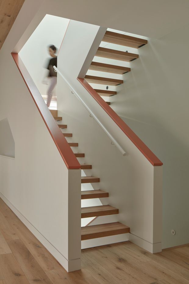 Interior photograph of a modern stairway with open-riser timber treads, white painted stringers and walls, and distinctive muted red handrail and trim accents. A mirrored or reflective panel beside the stairs reflects a blurred person, while exposed timber flooring and open stair construction create a light, contemporary feel.