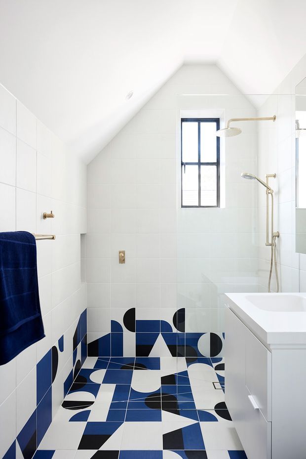 Bright contemporary bathroom with a sloped, vaulted ceiling and white tiled walls. The room features a freestanding white bathtub, glass shower enclosure with brushed-gold fixtures, a narrow black-framed window, mirrored cabinetry, and a striking geometric floor and lower-wall tile pattern in navy blue, white, and black. A dark blue towel adds contrast.
