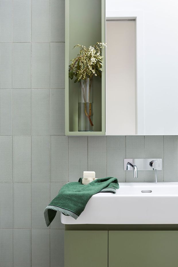 A close-up view of a refined contemporary bathroom featuring pale green small-format tiled walls, a white wall-mounted basin with exposed chrome tapware, and matching pale green vanity cabinetry. A recessed display niche with a vase of foliage and a white mirrored cabinet add decorative and functional detail. A dark green towel provides the main accent.