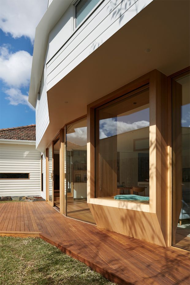 Exterior view of a contemporary residential extension with a warm timber deck, large timber-framed glazed doors and windows, and white horizontal weatherboard cladding. The covered upper volume projects over the deck, creating a sheltered outdoor threshold. An adjacent weatherboard structure with a tiled roof is visible, along with a small portion of lawn and blue sky.