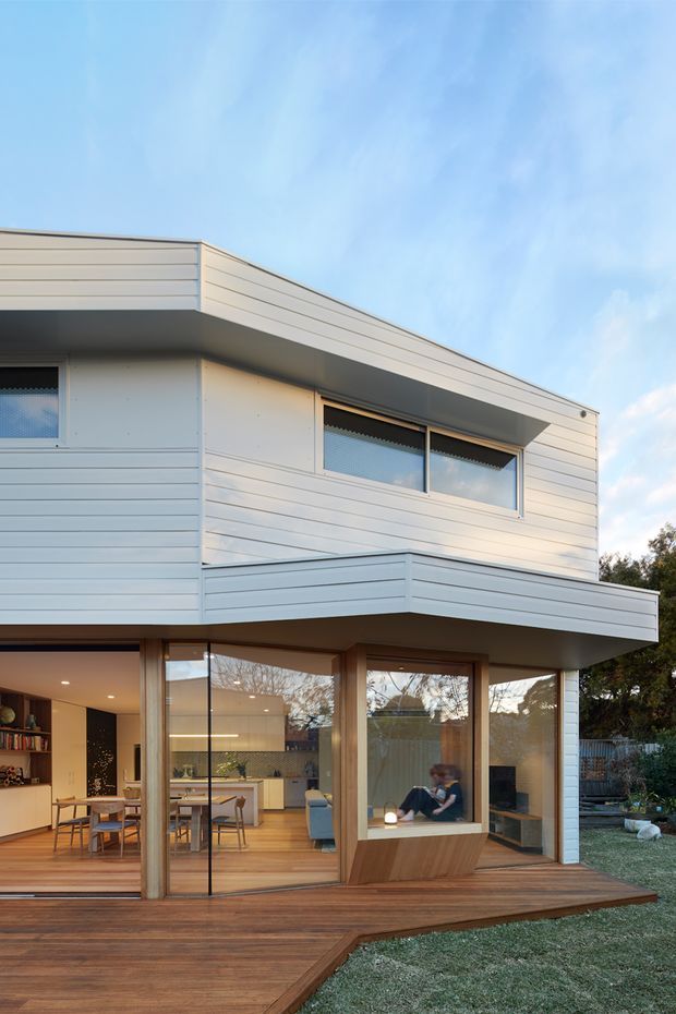 Exterior view of a contemporary two-storey residence with pale horizontal weatherboard cladding, a low-pitched roofline, projecting upper-level balcony, and extensive ground-floor glazing opening onto a timber deck. A warm open-plan kitchen and dining area is visible through the glass, with a landscaped yard surrounding the deck.