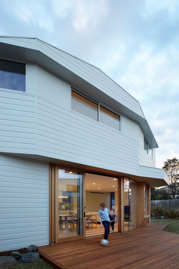 Exterior view of a contemporary multi-storey home with a steeply pitched roof and extensive white horizontal weatherboard cladding. Large timber-framed glazed doors open from the ground floor onto a timber deck, revealing a warm, light-filled dining and living area inside. The upper level has horizontal windows beneath deep roof eaves, and landscaped rocks and fencing sit around the deck.