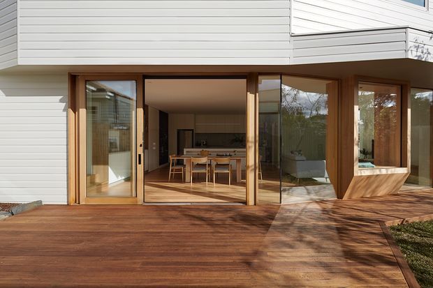 Exterior view of a contemporary home with white horizontal cladding, warm timber-framed full-height glazing, and a broad timber deck. Open bi-fold or sliding doors reveal a dining area and kitchen inside, while an angled glazed bay projects from the facade.