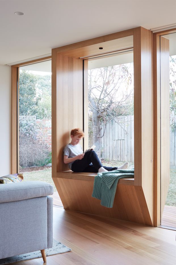 Bright contemporary interior featuring a built-in timber window seat integrated into a large glazed wall. Warm wood framing and flooring surround expansive windows overlooking a garden, while a grey upholstered sofa and teal throw add soft furnishings. A child is seated reading in the deep window nook.