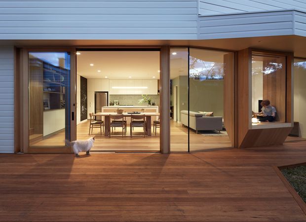 Contemporary residential exterior viewed from a broad timber deck, with expansive floor-to-ceiling glazing opening onto a combined dining and kitchen area. The home has pale horizontal cladding, warm timber-framed doors and windows, timber decking, and a projecting upper level. A living area and seated person are visible through the glazing, with a cat on the deck.