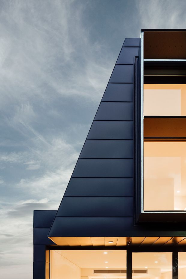 Close-up exterior view of a contemporary multi-storey building, featuring a sharply sloping dark metal-clad roof or wall plane, crisp black window and door frames, and large illuminated glazed openings. Warm interior lighting contrasts with the cool blue-grey sky and dark facade.