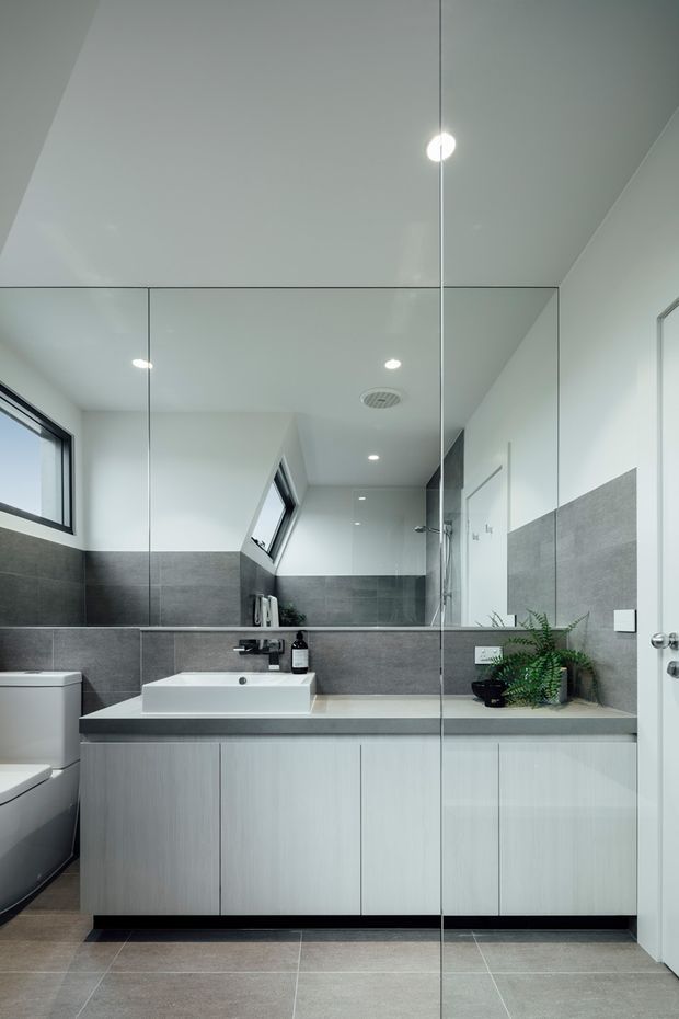 Contemporary bathroom with a floating light-wood vanity, white rectangular basin, dark grey benchtop, large grey wall and floor tiles, frameless glass shower screen, mirrored wall, recessed ceiling lights, skylight-style windows, and a toilet partially visible at left. A small fern adds greenery to the otherwise monochromatic space.