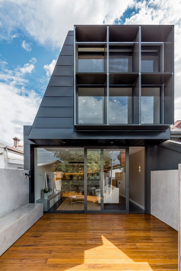 Photograph of a contemporary multi-storey rear facade and outdoor timber deck. The dark metal-clad upper level projects outward with a strongly gridded arrangement of floor-to-ceiling windows and deep boxed reveals. The ground level has broad glazed sliding doors opening onto the deck, with a modern living area visible inside. A neighbouring traditional roofline is partially visible at the side.