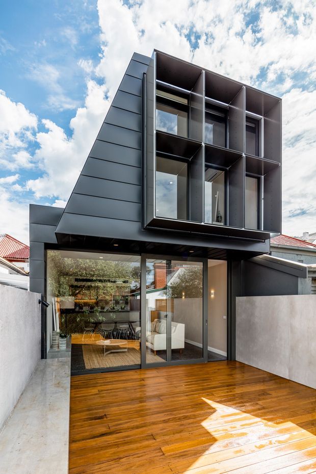 Exterior view of a compact contemporary multi-storey urban home extension. The dark metal-clad upper level projects over a glazed ground floor, with a dramatic sloping side wall and deep framed window bays. A timber deck occupies the foreground, while neighbouring traditional homes with red roofs are visible around the property.