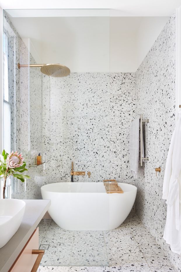 Bright contemporary bathroom featuring a freestanding white bathtub, wall-mounted brass tapware, a large rainfall shower behind clear glass, and extensive white terrazzo-style wall and floor tiling with dark stone flecks. A partial white basin and timber-toned vanity are visible in the foreground, with a heated towel rail and soft natural light from the window.