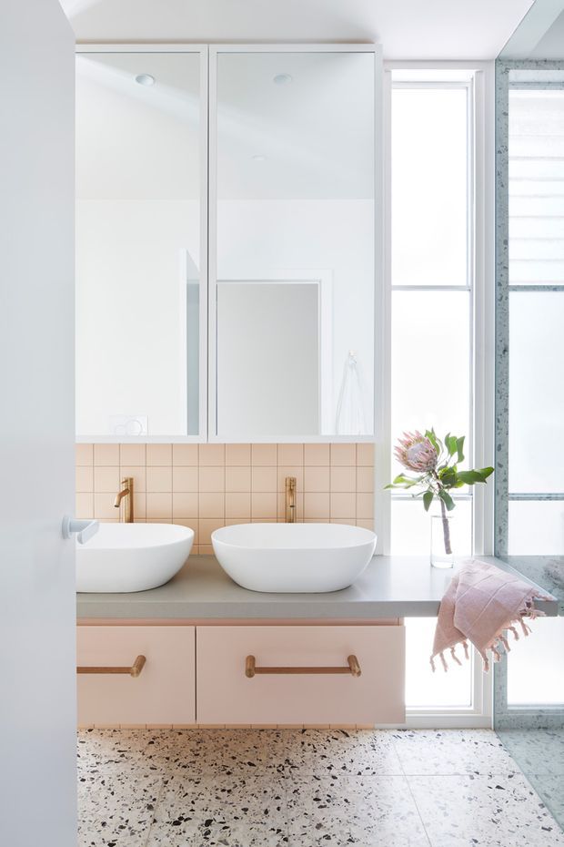 Bright contemporary bathroom featuring a double vanity with two white vessel basins, pale timber cabinetry with brass pulls, a grey benchtop, and a soft blush-pink square-tile splashback. Large mirrored wall cabinets reflect the room, while a tall window and glazed shower enclosure bring in abundant natural light. The floor is finished in terrazzo with dark aggregate.