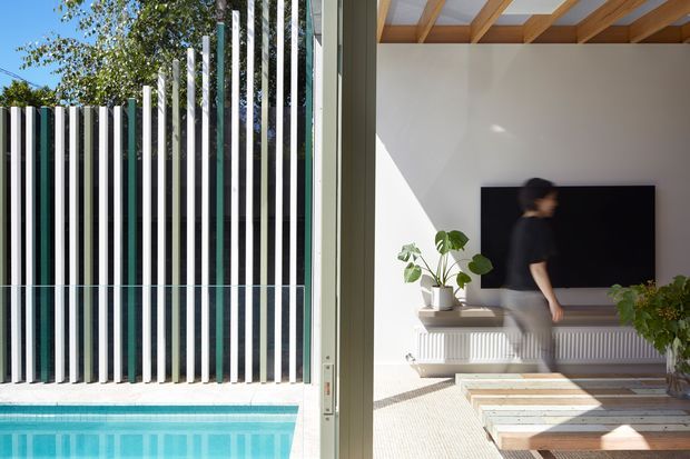 A split view through open glazing shows a bright living room beside a swimming pool. The interior features a wall-mounted television, low light-coloured joinery or a radiator cabinet, a pale textured floor, indoor plants, and a timber-framed exposed-beam ceiling. Outside, the pool is bordered by a distinctive vertical slatted screen in alternating white and dark tones.