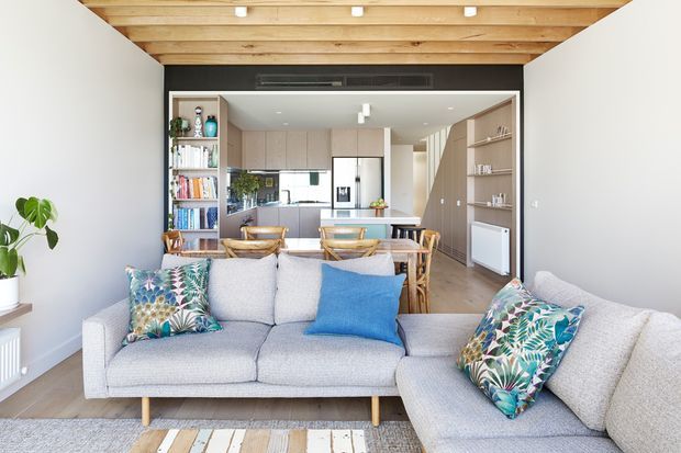Bright open-plan living and dining area leading into a contemporary kitchen. A large light-grey sectional sofa with blue and patterned cushions occupies the foreground, while a timber dining table and chairs sit behind it. The kitchen has pale timber cabinetry, integrated appliances, open shelving, and a dark feature wall. Exposed timber ceiling beams add warmth and architectural character, with light timber flooring and white walls creating an airy, inviting interior.