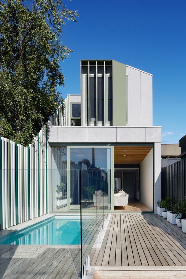 Exterior architectural photograph of a contemporary multi-storey residence with a rectilinear white and pale green facade, tall black-framed vertical glazing, and a recessed timber-lined entry or outdoor living zone. A narrow turquoise swimming pool runs alongside a timber deck with clear glass balustrading, surrounded by privacy fencing and planting. Bright blue sky and a mature tree frame the urban residential setting.