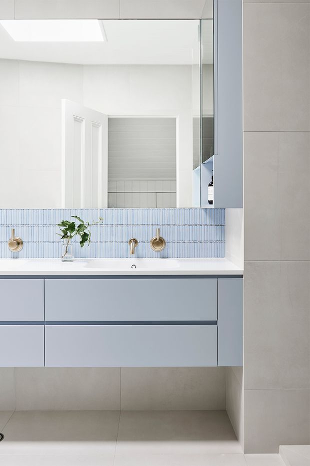 Bright contemporary bathroom vanity with a wall-mounted pale blue-grey cabinet, white benchtop, integrated basin, brass wall-mounted taps, and a narrow blue-and-white vertical mosaic tile splashback. Large mirror-fronted storage, light grey floor and wall tiles, and a skylight create a clean, airy appearance.
