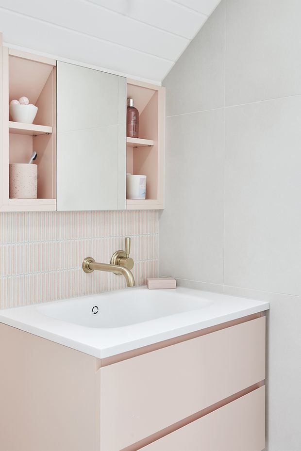 A compact, softly styled bathroom vanity with a white integrated basin, pale blush-pink drawer cabinetry, brushed brass wall-mounted tapware, and a mirrored medicine cabinet with open pink-lined shelving. The backsplash features narrow vertical pastel tiles, while large pale grey wall tiles and a white panelled ceiling create a clean, bright finish.