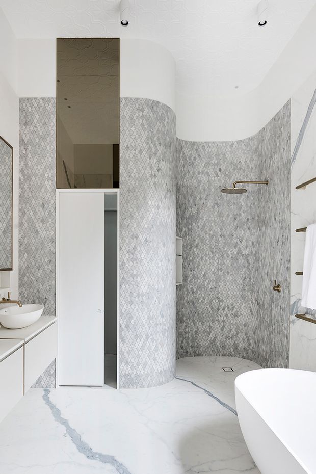 A bright, high-end contemporary bathroom with a freestanding white bathtub, wall-mounted brass shower fittings, a curved shower enclosure lined with small diamond-patterned grey and white tiles, and a white vanity with vessel basin. The floor and lower wall surfaces feature white marble-look stone with pronounced grey veining. A mirrored recessed cabinet and curved white ceiling details contribute to the refined, sculptural design.