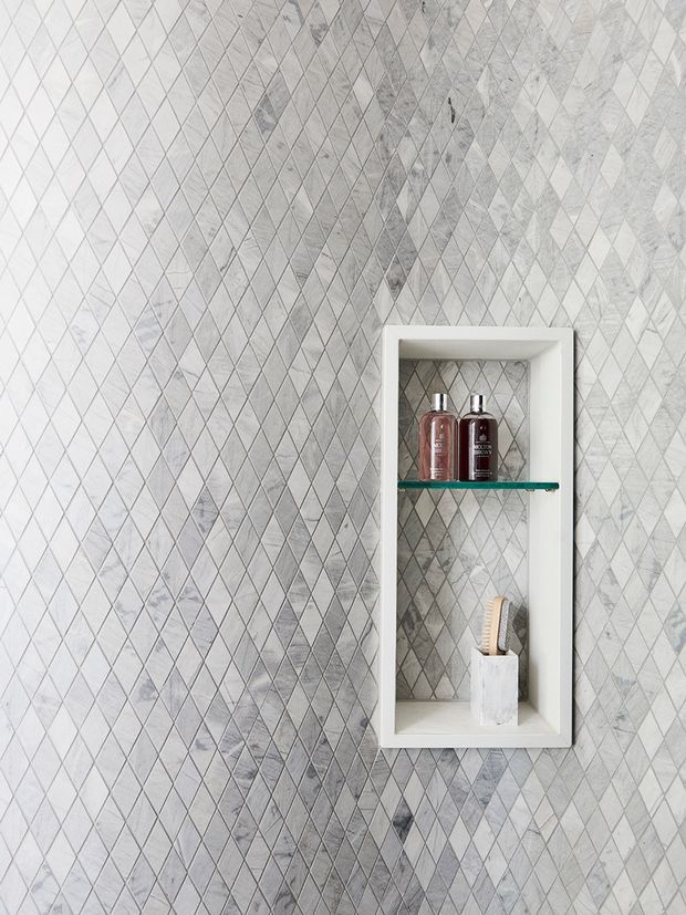 Close-up of a bathroom wall finished in a dense diamond-pattern mosaic of pale grey and white stone-look tiles. A recessed white-framed niche contains a glass shelf with toiletry bottles and a small holder, creating a clean, refined built-in storage detail.