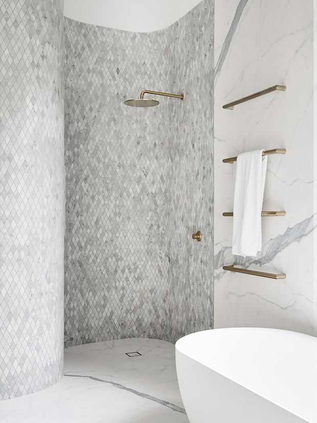 A luxurious contemporary bathroom featuring a curved walk-in shower clad in small grey diamond-patterned tiles, large white marble-look wall and floor surfaces with prominent veining, and a freestanding white bathtub. Brushed brass shower hardware and multiple towel rails add warm metallic accents. The space is bright, refined, and sculptural, with curved architectural forms and a restrained neutral palette.