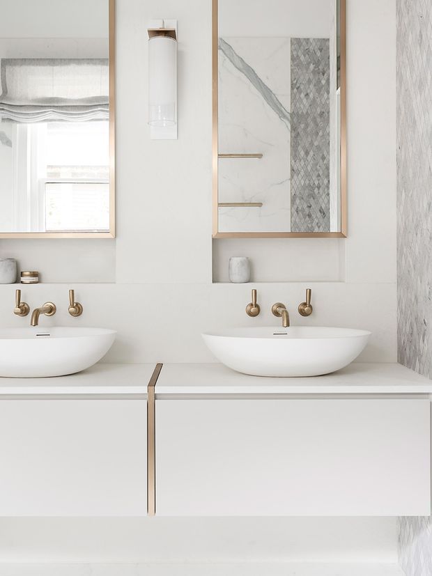 A bright, refined bathroom vanity featuring two white vessel basins on a floating white joinery unit. The wall-mounted basins are paired with brass/gold wall-mounted taps and framed mirrored cabinets. One cabinet includes marble-look slab panels and a vertically patterned tile feature, while the right wall is clad in a pale marble-look textured surface. A slim wall light is mounted between the mirrors.