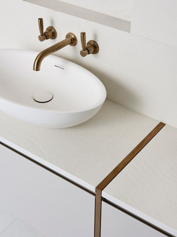 Close-up of a minimalist bathroom vanity featuring a white vessel basin, wall-mounted bronze-gold tapware, a pale textured benchtop, and light cabinetry with a contrasting dark timber accent edge.