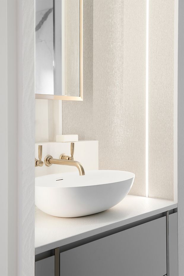 Close-up of a refined contemporary bathroom vanity. A white vessel basin sits on a pale stone or engineered-stone benchtop above dark grey fixed cabinetry. The wall features light textured vertical panelling, with a slim framed mirror and brushed brass/gold wall-mounted tapware. The space is bright, minimal, and softly neutral.