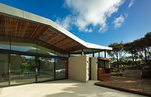 Low-slung contemporary building exterior with broad roof overhangs, a timber slatted soffit, extensive dark-framed glazing, pale masonry walls, and a timber deck. The landscaped setting includes stepped planting beds, while a clear blue sky and trees surround the structure.