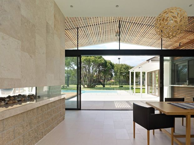 A refined open-plan living and dining area opening through full-height black-framed glazing to a pool terrace and landscaped grounds. The interior features a large stone fireplace, pale tiled flooring, a timber dining table, and a striking curved slatted timber ceiling with a sculptural pendant light.