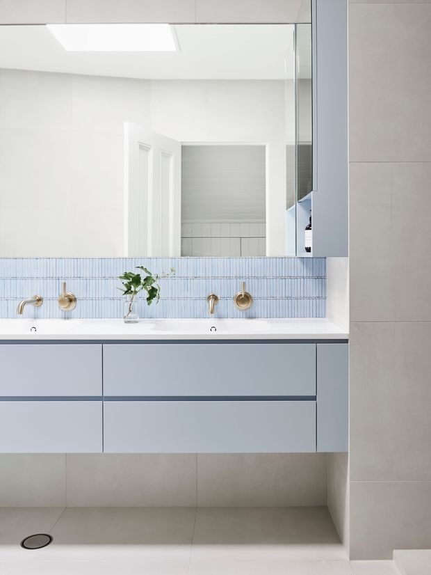 Bright contemporary bathroom with a wide floating double vanity in pale blue-grey cabinetry, white basins, brushed brass wall-mounted taps, and a narrow blue vertically ridged tile splashback. Large-format light grey wall and floor tiles create a clean, minimalist finish. A large mirror reflects an open white door and tiled adjoining area.