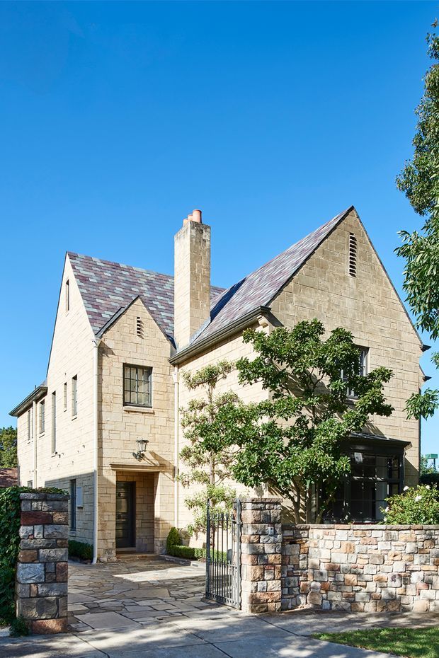 Exterior view of a substantial stone-clad house with multiple steep gabled roof sections, a prominent masonry chimney, small leaded-style windows, and a landscaped front entry. A low rough-stone boundary wall and metal pedestrian gate frame the paved approach. The facade uses pale beige masonry with dark trim and a patterned muted slate roof, under a clear blue sky.