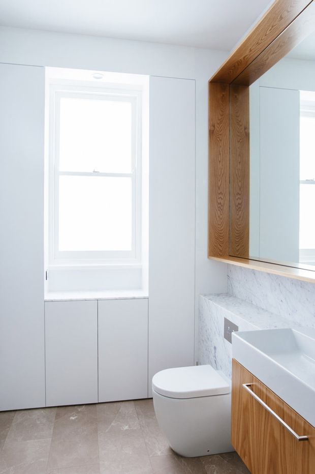 Bright contemporary bathroom featuring a wall-hung toilet, white integrated basin, large mirrored cabinetry, white marble-look wall cladding, warm timber joinery, pale stone-look flooring, and a white-framed window.
