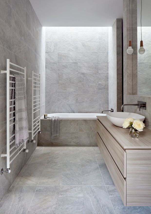 A compact contemporary bathroom with grey stone-effect wall and floor tiles, a built-in bathtub at the rear, white heated towel rails, and a floating timber-look vanity with a white vessel basin. A large mirror and two pendant lights sit above the vanity, creating a clean, softly lit interior.