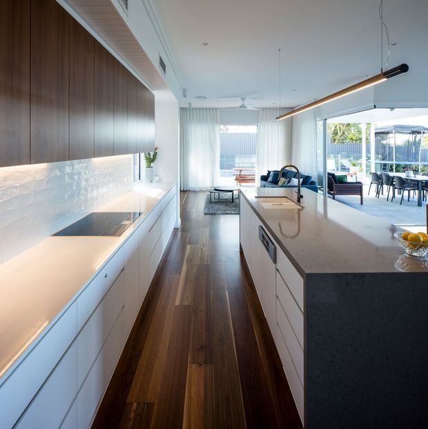 Bright, contemporary open-plan kitchen with a long central island, dark timber flooring, white flat-panel cabinetry, dark timber wall joinery, a glossy pale splashback, and a light stone benchtop. The kitchen opens to a sunlit living and dining area with full-height glazing and an outdoor entertaining space beyond. A linear pendant light and concealed ventilation contribute to the streamlined design.