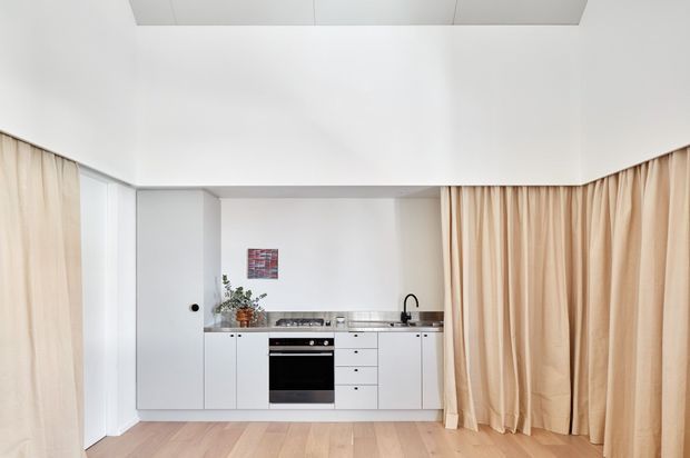 Compact open-plan kitchen with white flat-panel cabinetry, stainless-steel benchtop, integrated black oven and cooktop, and a black tap. Full-height beige curtains frame the room, while pale timber flooring and a white ceiling create a bright, minimal interior. A small artwork and potted plant add limited decoration.