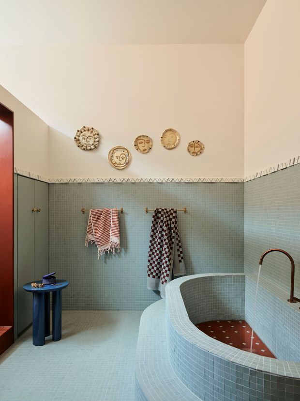 A distinctive tiled bathroom with a large curved built-in bathtub clad in small pale blue-green mosaic tiles. Matching mosaic wall and floor tiles are topped by a decorative geometric border and warm off-white painted walls. Dark green built-in cabinetry, brass hooks, patterned towels, sculptural wall plates, and a deep blue side table add colour and character. A copper-toned arched tap and reddish patterned tub interior provide focal accents.
