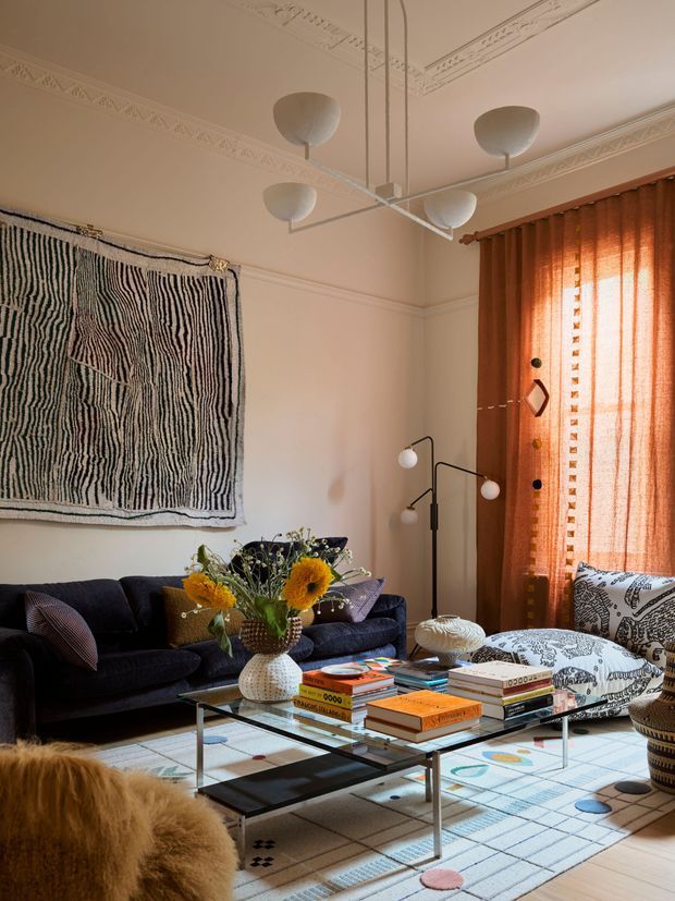 A richly layered living room with a dark navy sofa, patterned armchairs, a glass and metal coffee table stacked with books, a colourful geometric rug, sculptural floor lamps, and a large abstract textile wall hanging. Warm rust-orange curtains filter sunlight beside an ornate high ceiling with decorative plaster cornices. The room has an eclectic, artistic, mid-century-influenced interior with strong pattern and texture.
