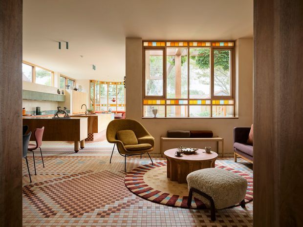 Open-plan mid-century-inspired interior combining a living room, dining area and kitchen. The room features extensive patterned tiled flooring, warm timber joinery, stained-glass window bands, large timber-framed windows, a sculptural olive armchair, a low round timber coffee table, and colourful upholstered seating. The space is warm, richly patterned and connected to greenery outside.