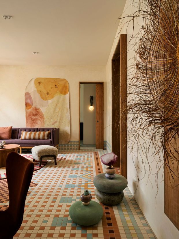 Eclectic, highly decorative living room with a warm cream palette, colourful geometric tiled flooring, a deep plum sofa, sculptural low tables, and a large abstract wall artwork. A wood-framed doorway and dark timber built-in shelving or panelling line the right side, while an oversized woven organic wall installation adds texture. The room has a playful, gallery-like character with layered colour, pattern, and sculptural furnishings.