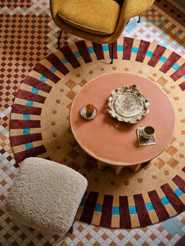 Top-down view of a richly layered seating area arranged around a low, round terracotta-coloured coffee table. A sculptural mustard upholstered armchair and a cream boucle lounge chair sit on a large circular rug with geometric burgundy, turquoise, beige and brown radial detailing. The table holds a decorative sculptural bowl or platter and coffee cups. Small multicoloured square floor tiles surround the rug, creating a highly patterned, warm and eclectic setting.
