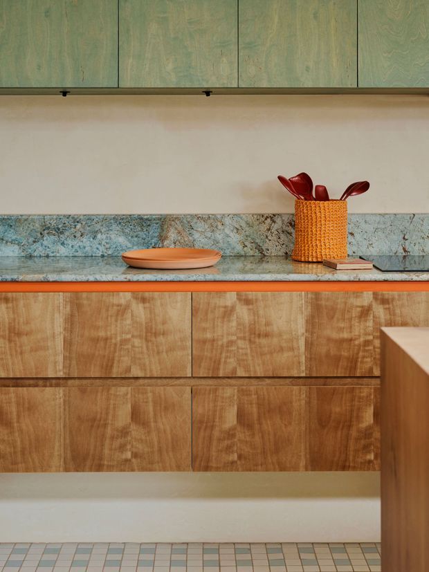 Close-up view of a richly detailed kitchen joinery composition, featuring warm timber-grain lower cabinetry, muted green timber-grain upper cabinets, a blue-green veined stone benchtop and backsplash, and a narrow orange accent trim. Decorative objects sit on the counter, while small square patterned floor tiles are visible below.
