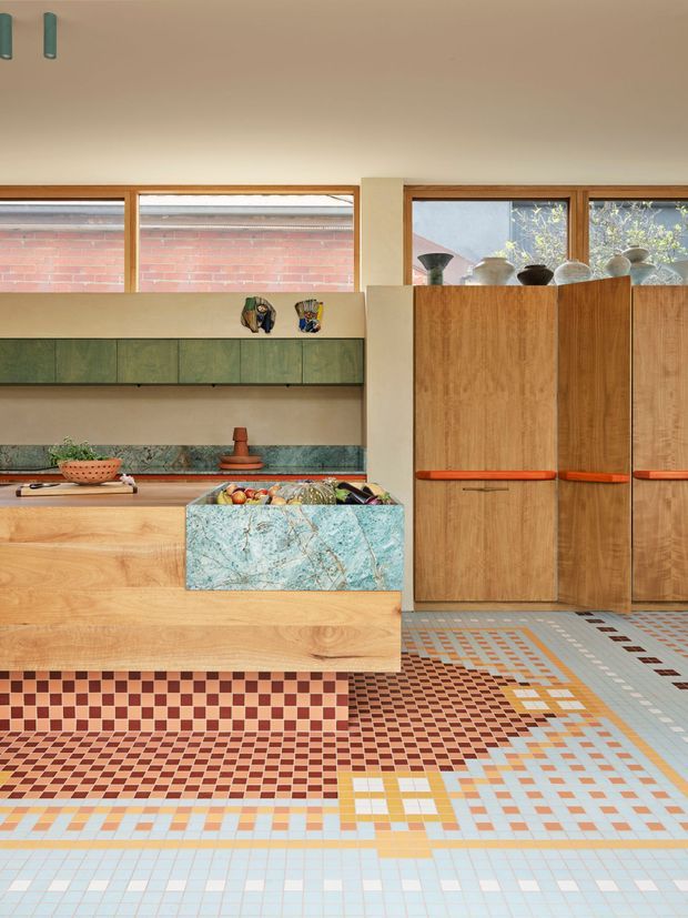 Colourful contemporary kitchen interior with extensive timber cabinetry and a long timber island, green built-in wall cabinets, a veined turquoise stone or marble island end, and a highly distinctive multicoloured geometric mosaic tile floor. Large timber-framed windows provide natural light, while sculptural pottery and small decorative objects sit above the cabinetry.