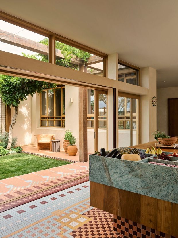 A sunlit, open-plan interior looking toward a landscaped courtyard through extensive timber-framed glazing. The foreground features a prominent fixed kitchen island with a richly patterned natural stone benchtop and timber cabinetry. The floor is covered in a highly decorative geometric mosaic tile pattern, while the courtyard includes lawn, mature planting, and a covered architectural structure. Warm plaster walls, exposed structural columns, and large glazed openings create a strong indoor–outdoor connection.