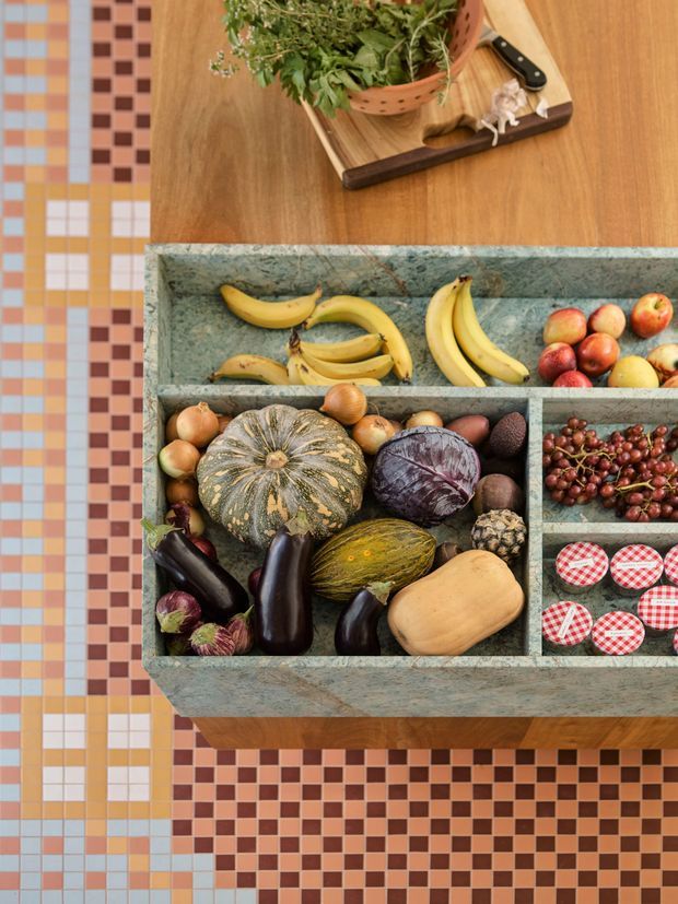 Top-down view of a colourful kitchen pantry or island storage arrangement. A timber worktop holds a wooden chopping board, knife, garlic, and leafy herbs, while a large integrated stone or terrazzo-like produce compartment below displays bananas, apples, onions, pumpkin, cabbage, eggplant, grapes, preserves, and other groceries. The surrounding floor or wall surface features a bold retro checkerboard mosaic tile pattern in terracotta, brown, cream, pale blue, and mustard tones.