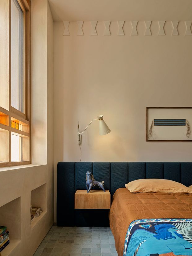 A warm, compact bedroom with a tall timber-framed multi-pane window, deep blue upholstered wall-to-wall headboard, and a rust-brown textured bedspread. A small timber bedside table holds a sculptural object, while a slim wall-mounted reading lamp and framed artwork add restrained character. The room features a pale beige plastered wall with a repeating raised geometric border near the ceiling, a tiled floor, and a built-in recessed shelf beneath the window.