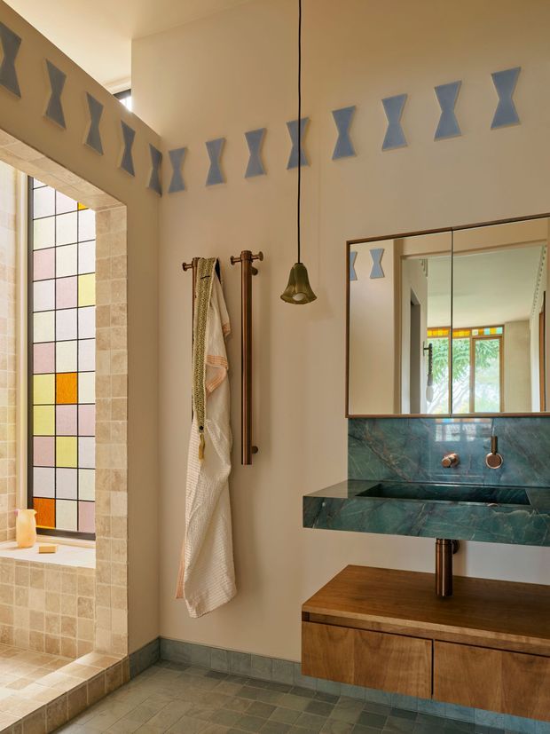 A warm, distinctive bathroom interior with a floating timber vanity, richly veined green stone basin and splashback, brass-toned wall-mounted basin fittings, and a large framed mirror. The room features a sculptural pendant light, a towel rail with hanging textiles, grey-green floor tiles, and a tall geometric stained-glass window framed in pale stone. Repeated blue-grey hourglass-shaped wall accents add a decorative architectural detail.