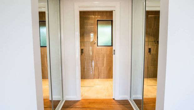 A compact bathroom viewed from a hallway or entry, with warm tan tiled walls and floor, a white-framed doorway, frosted glass window, and mirrored panels on both sides reflecting the space. Timber flooring is visible in the foreground.