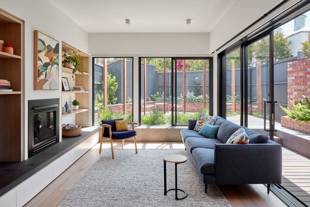 Bright contemporary living room with a dark blue sectional sofa, pale textured rug, timber built-in shelving and cabinetry, and an integrated fireplace surround. Extensive black-framed glazing opens the room visually to a planted courtyard and timber deck, while a low window seat runs along the rear windows. The room has a clean, warm modern character with abundant natural light.