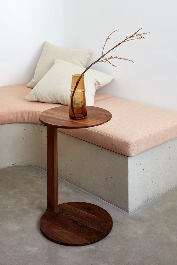 A softly styled minimalist interior vignette featuring a low concrete daybed or bench with a blush pink upholstered cushion and cream pillows. In the foreground is a dark timber pedestal side table with a round top and circular base, holding an amber glass vase with a bare branch. The space has white walls and a smooth grey concrete floor, creating a calm, restrained material palette.